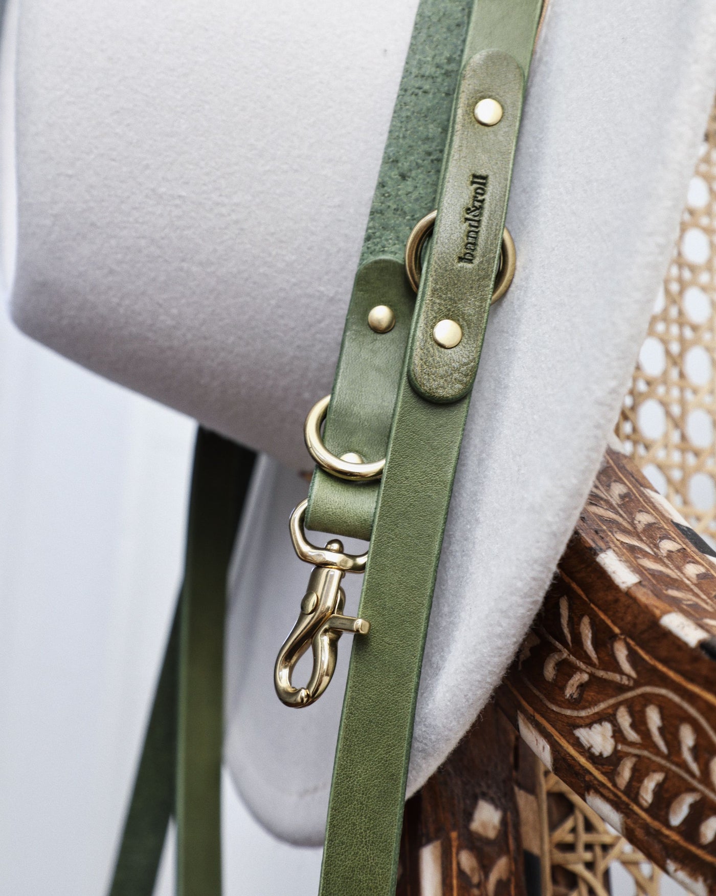 Close up of green leather crossbody dog leash and gold hardware hanging off hat on chair